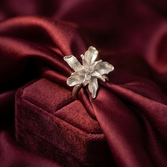 A silver flower-shaped ring displayed on top of a dark red velvet jewelry box, set against a draped dark red satin fabric background.
