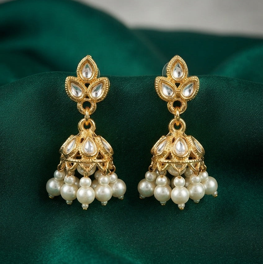 A pair of gold-plated Indian jhumka earrings adorned with clear stones and clusters of white faux pearls, displayed on a draped dark green fabric background.