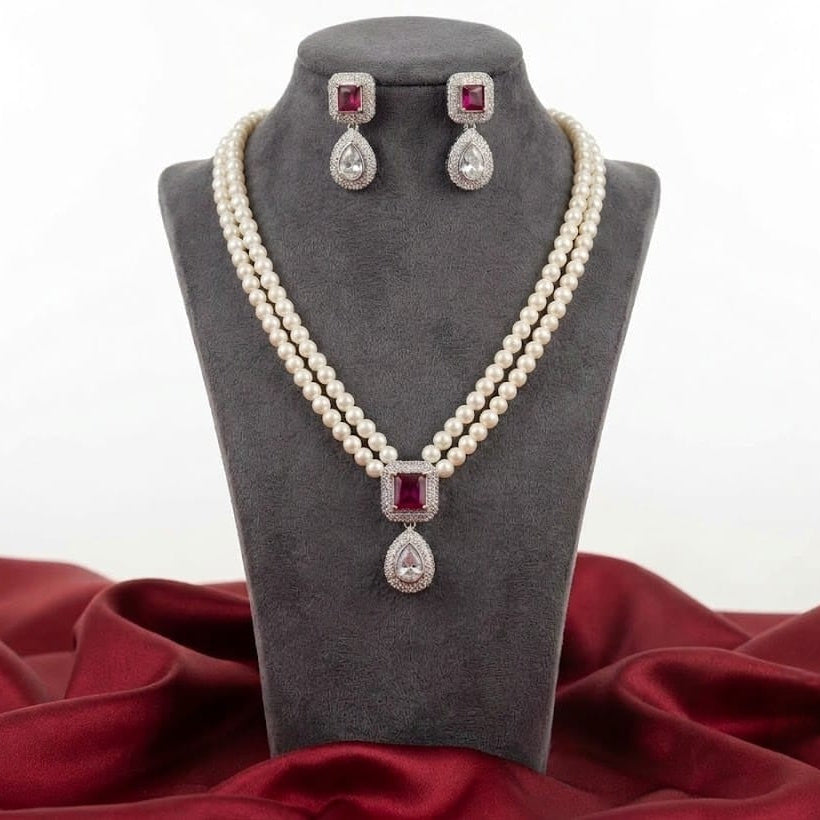 A pearl necklace and matching drop earrings displayed on a dark gray jewelry bust. The necklace and earrings feature square red gemstones in silver-toned settings with teardrop-shaped clear stones below.