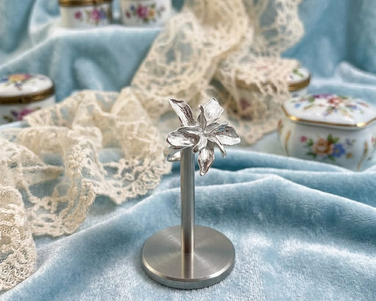Silver flower-shaped object on a stand with lace and decorative boxes in the background.