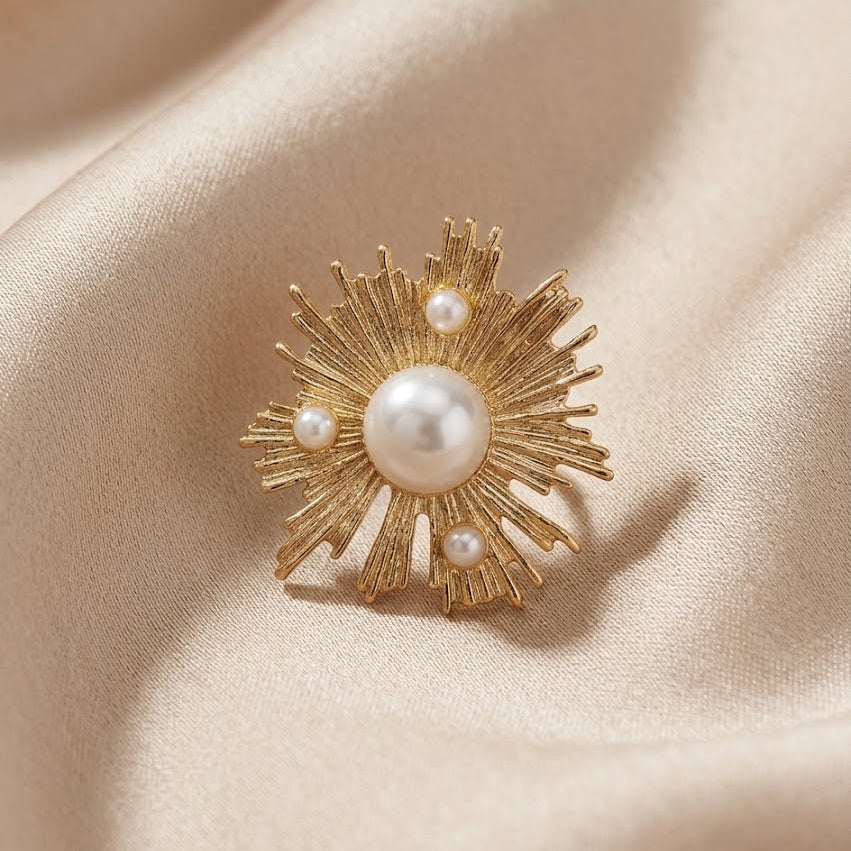 A gold sunburst brooch with a central white pearl and three smaller pearls, displayed on a draped beige satin fabric.