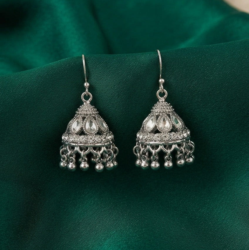A pair of silver-toned Indian jhumka earrings with intricate filigree work and dangling bells, displayed on a draped emerald green satin fabric.