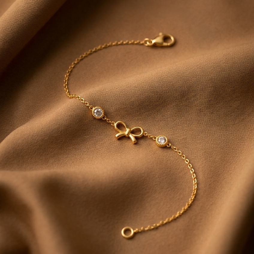 A delicate gold chain bracelet with a small bow charm and two small clear gemstone accents, displayed on draped brown fabric.
