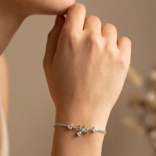 A close-up of a person's hand and wrist wearing a delicate silver bracelet with a small floral or butterfly-like charm set with clear stones, against a soft beige background.