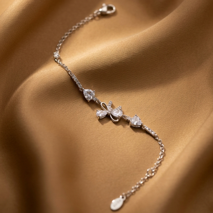 A delicate silver chain bracelet with a small bow charm set with clear stones, displayed on draped tan satin fabric.