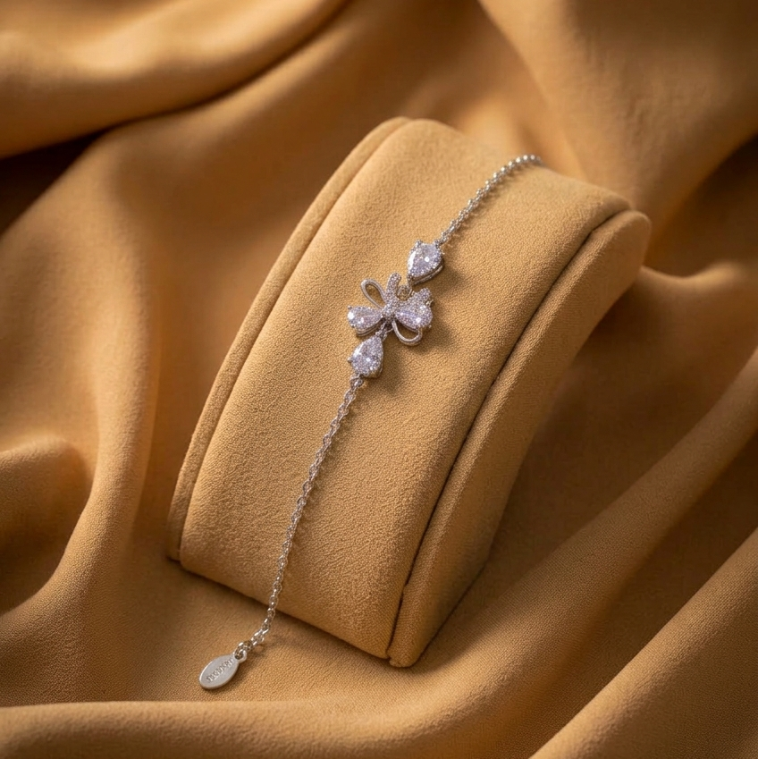 A delicate silver-toned chain bracelet with a floral motif and clear stones, displayed on a matching cream-colored jewelry cushion against a draped tan fabric background.
