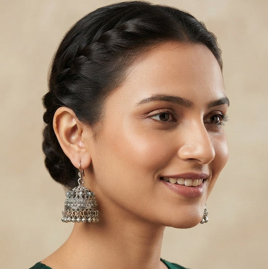 A woman with dark hair styled in a braided bun is shown in a close-up portrait, wearing a deep green embroidered top and a large silver-toned jhumka earring. The background is a soft, warm beige.