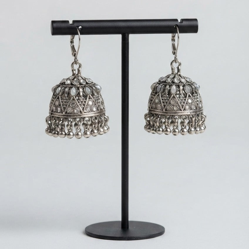 A pair of silver-toned Indian jhumka earrings with intricate filigree detailing and dangling bell-shaped charms, displayed on a black metal stand against a light gray background.