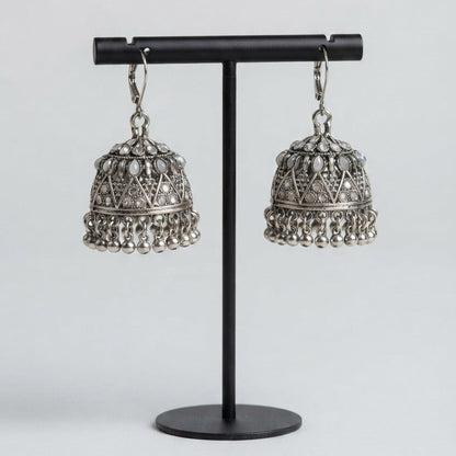A pair of silver-toned Indian jhumka earrings with intricate filigree detailing and dangling bell-shaped charms, displayed on a black metal stand against a light gray background.