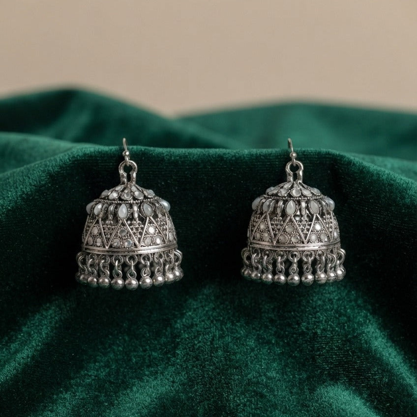 A pair of silver-toned Indian jhumka earrings with intricate geometric filigree and dangling bead borders, displayed on a draped dark green velvet fabric.