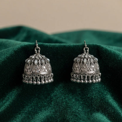 A pair of silver-toned Indian jhumka earrings with intricate geometric filigree and dangling bead borders, displayed on a draped dark green velvet fabric.