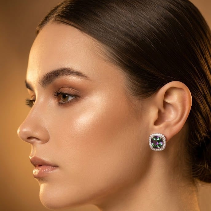 A close-up side profile of a woman wearing a square stud earring with a green gemstone surrounded by a silver-toned halo setting.