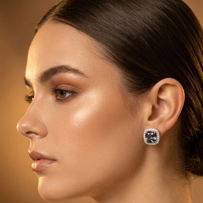 A close-up side profile of a woman wearing a square stud earring with a green gemstone surrounded by a silver-toned halo setting.