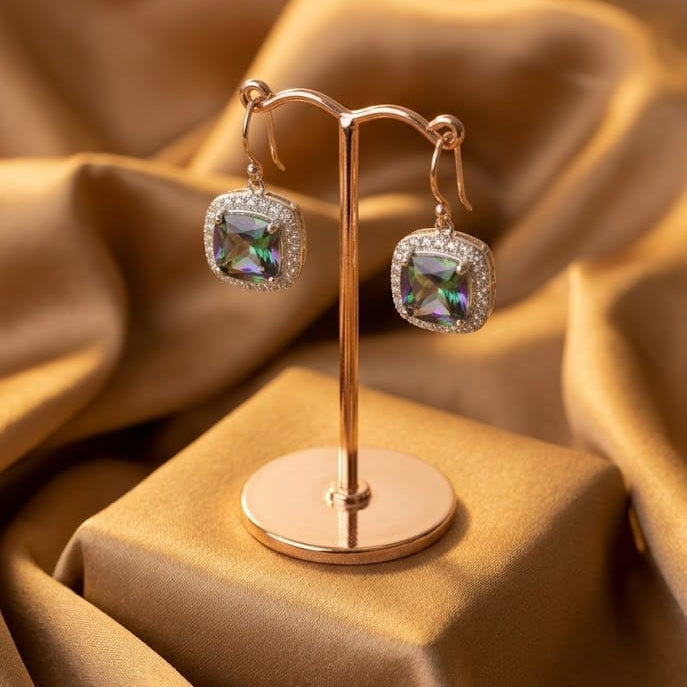 Square iridescent gemstone drop earrings displayed on a rose gold stand, set against a draped gold satin fabric background.