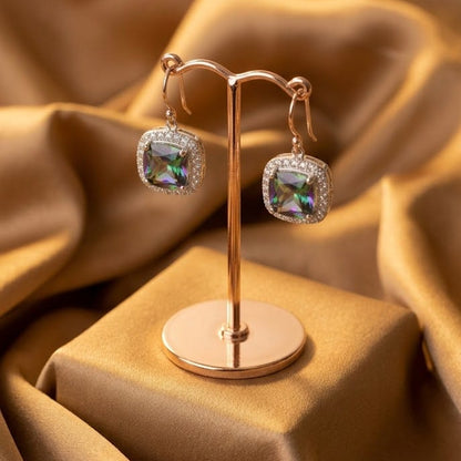 Square iridescent gemstone drop earrings displayed on a rose gold stand, set against a draped gold satin fabric background.