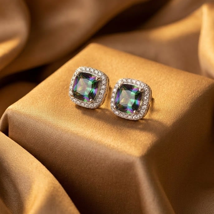 A pair of square stud earrings with iridescent green-blue center stones and clear halo stones, displayed on a gold satin block against a draped gold satin background.