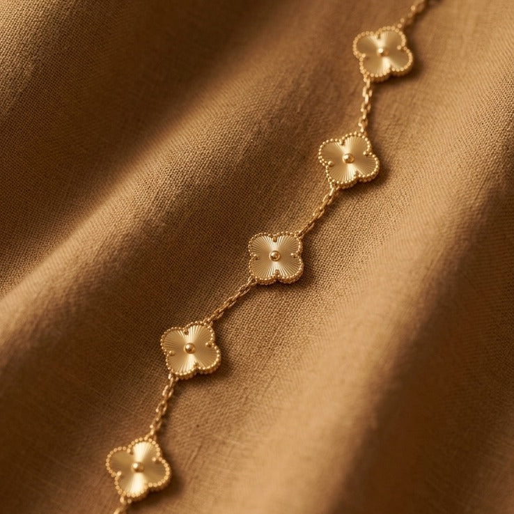 A delicate gold chain bracelet featuring five small clover-shaped charms, displayed diagonally on a draped brown fabric background.