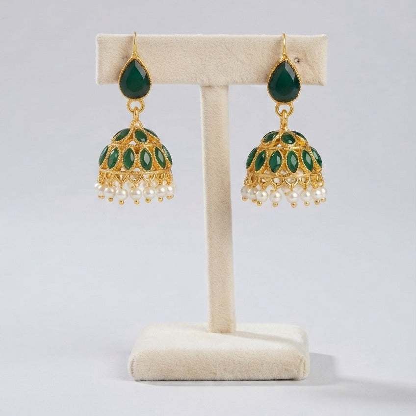 A pair of gold-plated Indian jhumka earrings with green enamel detailing and dangling white pearl beads, displayed on a cream-colored jewelry stand against a light gray background.