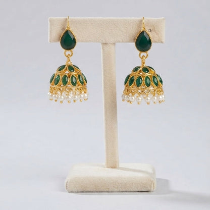 A pair of gold-plated Indian jhumka earrings with green enamel detailing and dangling white pearl beads, displayed on a cream-colored jewelry stand against a light gray background.