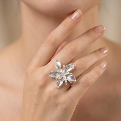 A silver flower-shaped ring worn on a woman's finger, shown in a close-up lifestyle shot against a soft beige background.