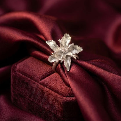 A silver flower-shaped ring displayed on top of a dark red velvet jewelry box, set against a draped dark red satin fabric background.