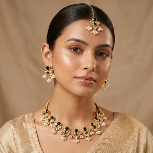 A woman wearing traditional Indian jewelry, including a gold-toned maang tikka, matching earrings, and a statement necklace with gold, black, and white elements, paired with a beige blouse against a neutral beige backdrop.