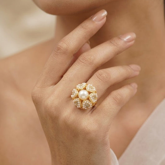 A close-up of a woman's hand wearing a gold ring with a central white pearl surrounded by small clear stones, against a soft beige background.