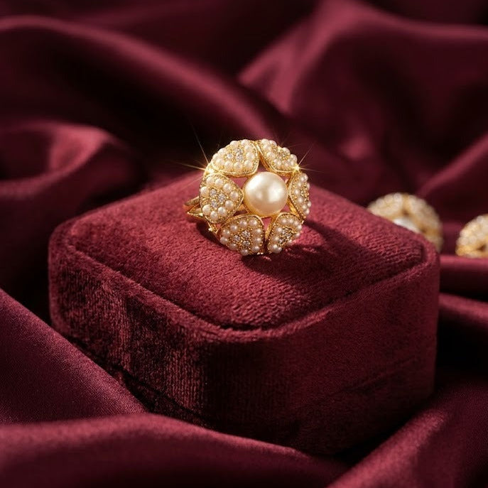 A gold ring with a central pearl and surrounding clear stones, displayed on a deep burgundy velvet box against a matching draped fabric background.