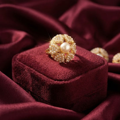 A gold ring with a central pearl and surrounding clear stones, displayed on a deep burgundy velvet box against a matching draped fabric background.