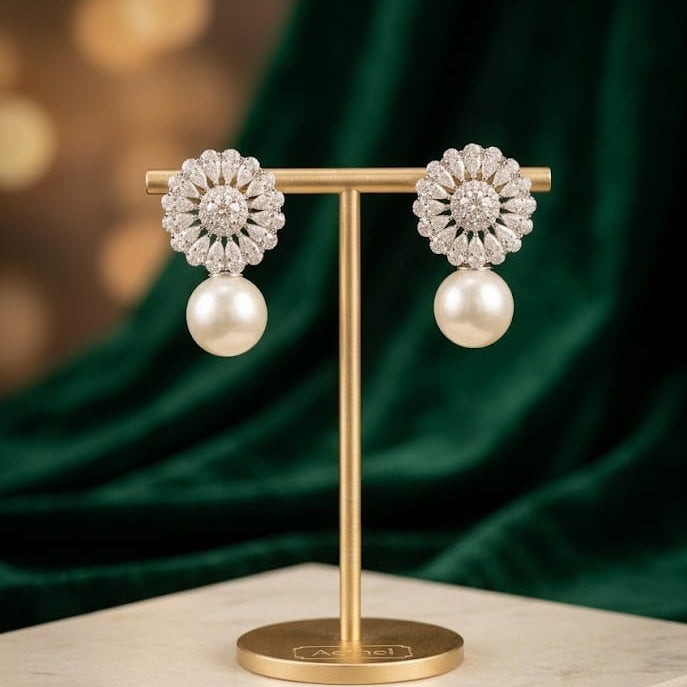 A pair of floral cluster earrings with white pearls hanging below, displayed on a gold T-stand against a dark green velvet backdrop and light marble surface.