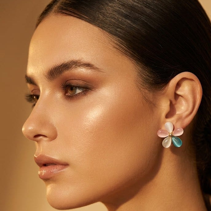 A close-up side profile of a woman wearing a delicate flower-shaped earring with pastel pink, white, and light blue petals, set against a warm beige background.