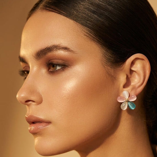 A close-up side profile of a woman wearing a delicate flower-shaped earring with pastel pink, white, and light blue petals, set against a warm beige background.