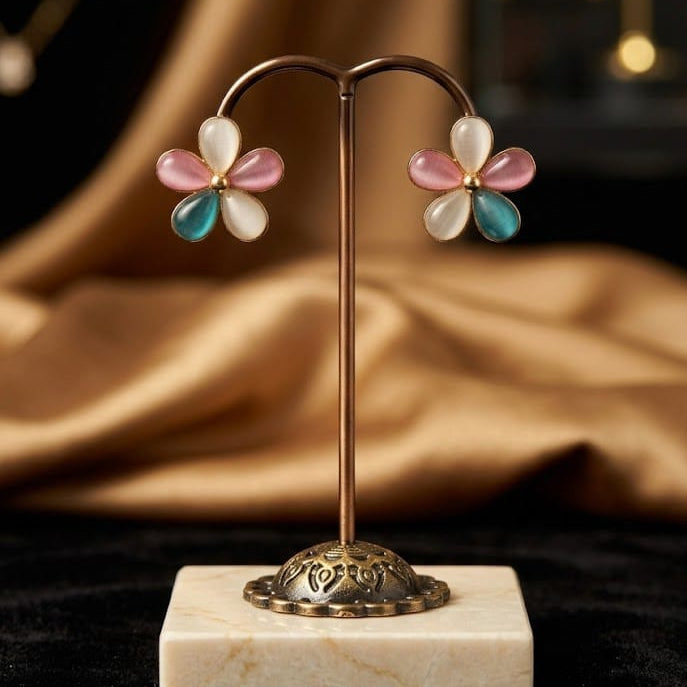 A pair of flower-shaped stud earrings displayed on a gold-tone stand with a white marble base. Each earring has five petals in white, pink, and teal, set against a dark background with draped gold fabric.