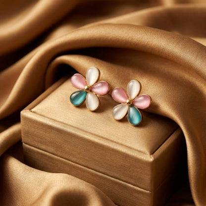A pair of flower-shaped stud earrings with pink, white, and teal enamel petals on a gold-tone setting, displayed on a small gold jewelry box against draped satin fabric.