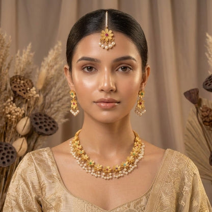 A woman wearing a gold and pearl traditional Indian jewelry set, including a maang tikka, earrings, and a necklace, paired with a beige embroidered blouse. She is framed by dried lotus pods and pampas grass against a neutral beige background.