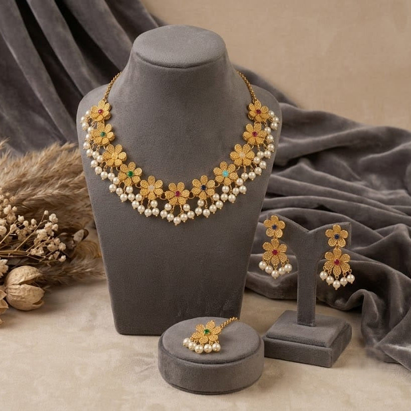 Gold floral jewelry set with white pearl-like beads and small green and orange accents, displayed on a gray mannequin bust with matching earrings and a ring holder on a beige surface with draped gray fabric and dried floral decor.