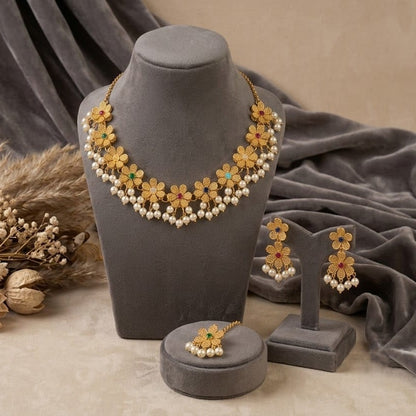 Gold floral jewelry set with white pearl-like beads and small green and orange accents, displayed on a gray mannequin bust with matching earrings and a ring holder on a beige surface with draped gray fabric and dried floral decor.