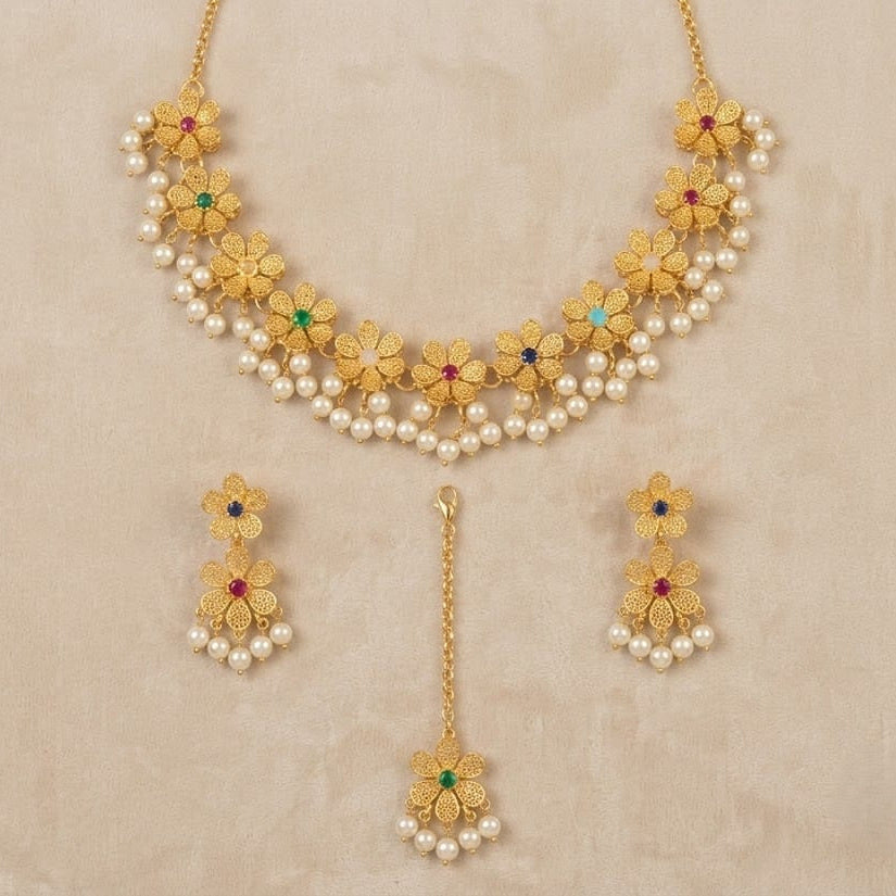 Gold-toned traditional jewelry set featuring a necklace, matching earrings, and a dangling pendant, decorated with faux pearls, gold floral motifs, and small red and green stone accents, displayed on a beige textured background.