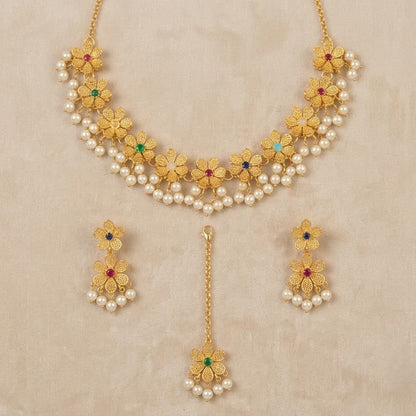Gold-toned traditional jewelry set featuring a necklace, matching earrings, and a dangling pendant, decorated with faux pearls, gold floral motifs, and small red and green stone accents, displayed on a beige textured background.