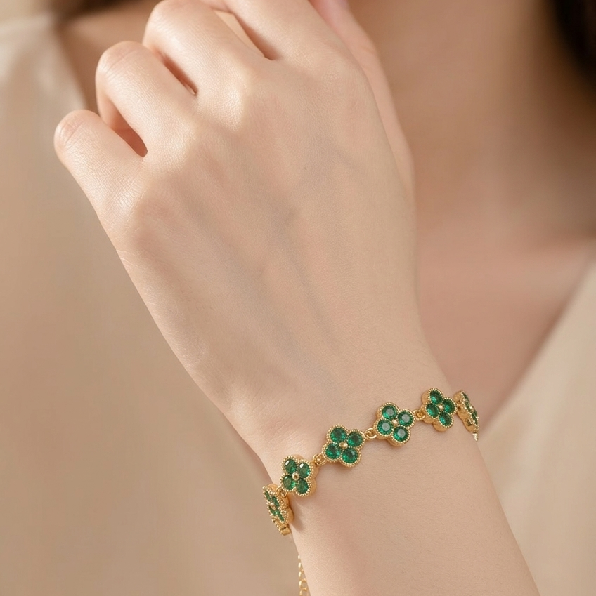 A close-up of a model's hand and wrist wearing a delicate gold bracelet adorned with five green clover motifs, set against a soft beige background.