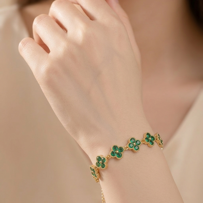 A close-up of a model's hand and wrist wearing a delicate gold bracelet adorned with five green clover motifs, set against a soft beige background.