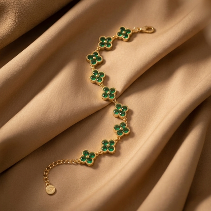 A gold-tone chain bracelet featuring seven clover-shaped links, each inlaid with vibrant green stones, displayed on draped beige satin fabric.
