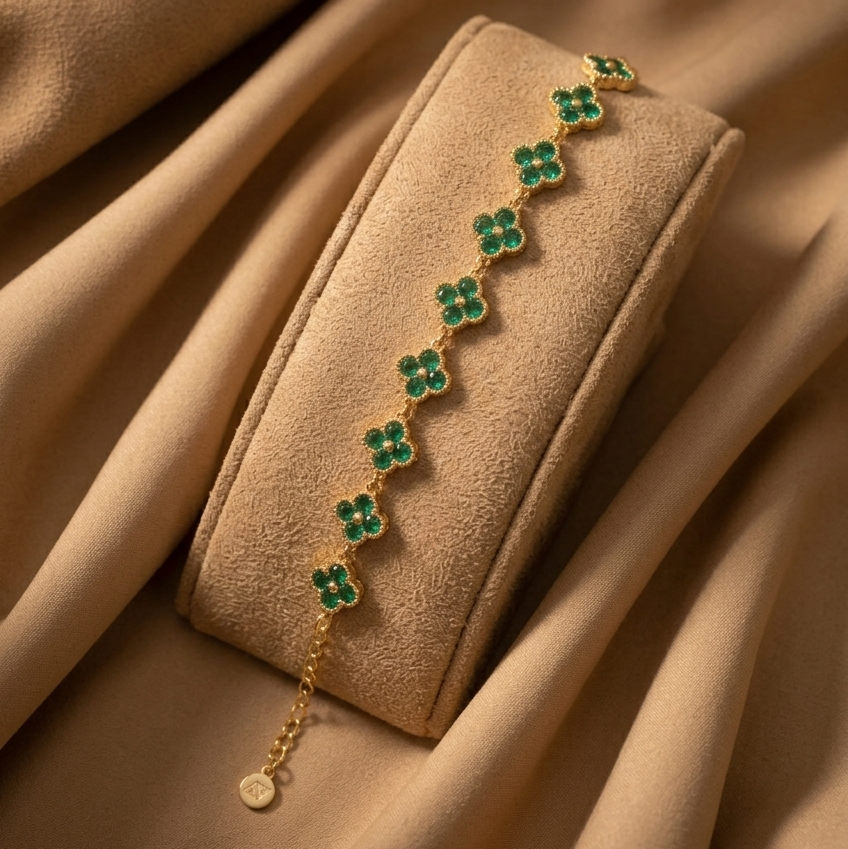 A gold chain bracelet with green clover motifs, displayed on a beige velvet jewelry roll against a draped beige fabric background.