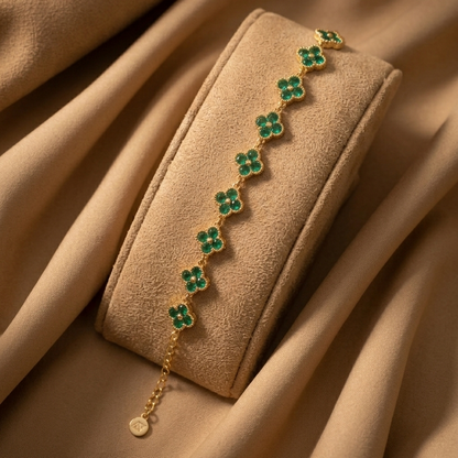 A gold chain bracelet with green clover motifs, displayed on a beige velvet jewelry roll against a draped beige fabric background.
