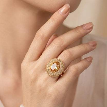 A close-up of a model's hand wearing a gold ring with a central pear-cut clear gemstone surrounded by a halo of small clear stones.