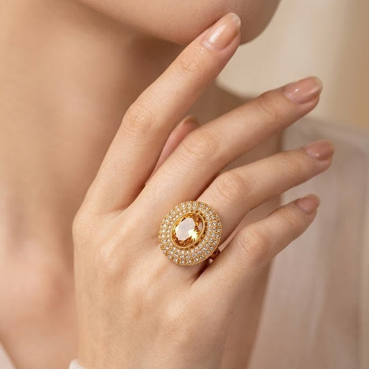 A close-up of a model's hand wearing a gold ring with a central pear-cut clear gemstone surrounded by a halo of small clear stones.