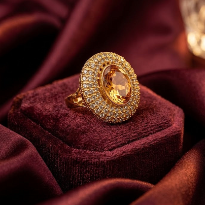 A gold ring with a large oval yellow gemstone surrounded by a halo of small clear stones, displayed on a burgundy velvet ring box against a draped burgundy fabric background.