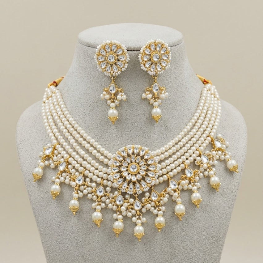 A traditional Indian jewelry set featuring a multi-strand pearl necklace with gold-plated embellishments, a matching round stud earring, and a central floral pendant, all displayed on a gray velvet jewelry bust against a light beige background.