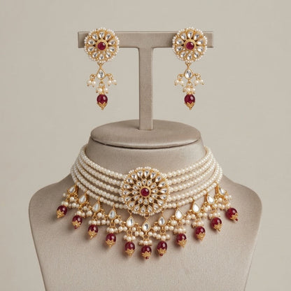 A traditional Indian jewelry set consisting of a multi-strand pearl choker necklace and matching drop earrings, embellished with gold-plated filigree, clear crystals, and deep red gemstone accents, displayed on a neutral beige jewelry stand.