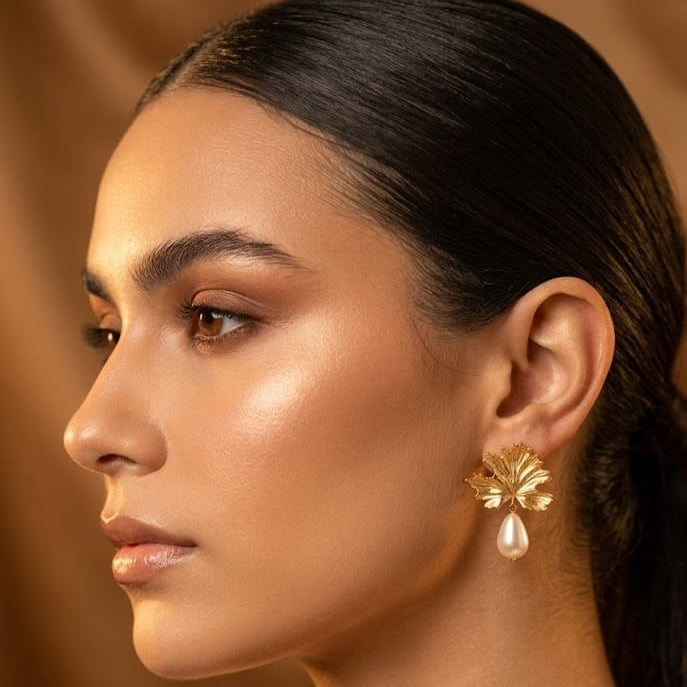 A close-up side profile of a woman with dark hair pulled back, wearing a gold flower-shaped earring with a small pearl drop, against a warm brown background.